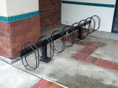 A black, durable steel, spiral-design bike rack is installed on a concrete and brick patio area next to a building wall